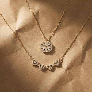 May include: Two gold necklaces with pendant charms. The top necklace features a four-leaf clover pendant with clear crystals. The bottom necklace has a series of heart-shaped pendants with clear crystals. Both necklaces are on a brown background.