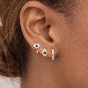 May include: Three gold earrings are displayed on an ear. The first is a small hoop with a row of clear stones. The second is a teardrop shape with a blue center stone and clear stones around it. The third is an eye shape with a blue center stone.