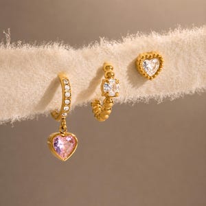May include: A set of three gold-toned earrings. One is a hoop with small clear stones and a dangling pink heart-shaped charm. The second is a beaded hoop with a central clear stone. The third is a small stud earring shaped like a heart with a clear stone.