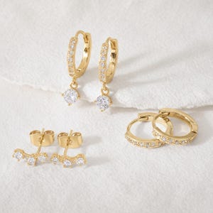 Puede incluir: Una colección de pendientes dorados. El conjunto incluye dos pares de pendientes de aro pequeños, uno con piedras transparentes colgantes y otro con piedras transparentes incrustadas. También se incluye un par de ear jackets con tres piedras transparentes.
