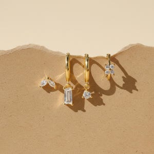 May include: A collection of gold-colored earrings with clear, sparkling gemstones. The earrings include a pair of small, leaf-shaped studs, two hoop earrings with rectangular and round gemstone pendants, and a small floral hoop earring. The earrings are displayed on a tan surface.