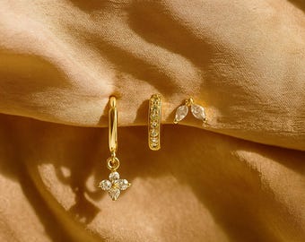 May include: Three gold-colored earrings on a beige background. One earring is a hoop with a dangling four-petal flower design. Another is a small hoop with embedded clear stones. The third is a stud with two marquise-shaped clear stones.