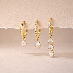 Puede incluir: Tres pares de pendientes de aro dorados. Un par tiene un solo colgante redondo de piedra preciosa transparente. El segundo par tiene un colgante de piedra preciosa transparente en forma de pera. El tercer par tiene tres colgantes de piedra preciosa transparente en forma de pera.