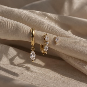 May include: A set of three gold-toned earrings consisting of one hoop earring with a dangling marquise-cut clear stone, one small hoop earring featuring three marquise-cut clear stones, and one single marquise-cut clear stone stud earring.