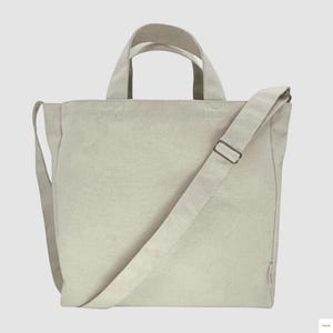 May include: A light beige canvas tote bag with two short handles and a long, adjustable shoulder strap. The bag has a rectangular shape and a simple, minimalist design, suitable for carrying various items.