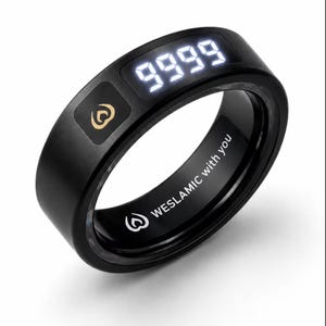 May include: A black smart ring featuring a digital display showing the number 9999 in white light. The outer surface includes a gold-colored flame-like logo. The inner band is engraved with the text WESLAMIC with you and a small flame icon.