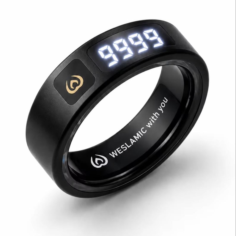 May include: A black smart ring featuring a digital display showing the number 9999 in white light. The outer surface includes a gold-colored flame-like logo. The inner band is engraved with the text WESLAMIC with you and a small flame icon.