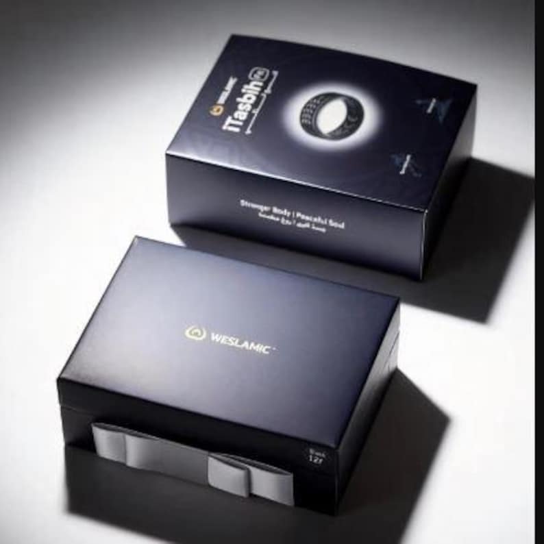 May include: Two dark blue rectangular product boxes. The top box features the text iTasbih, an image of a black ring, and the phrase Stronger Body | Peaceful Soul. The bottom box displays the WESLAMIC logo and a grey ribbon pull tab on the front.