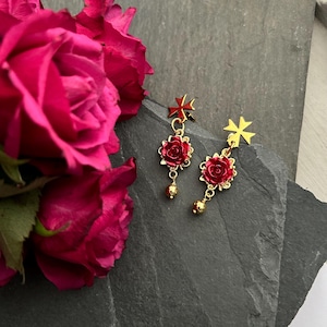 May include: A pair of gold earrings with a red rose charm and a gold cross charm. The earrings are dangling from a gold chain.