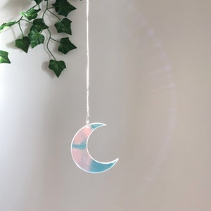 May include: A silver chain hangs from the ceiling with a crescent moon-shaped ornament at the end. The ornament is made of iridescent acrylic and reflects light in shades of pink and blue.
