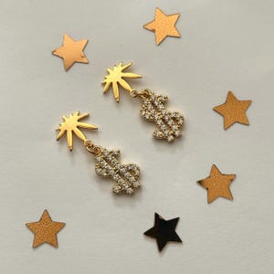 May include: Gold-colored earrings with a sunburst design and a dangling dollar sign encrusted with small, clear stones. The earrings are set against a white background with scattered gold and black star-shaped confetti.
