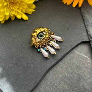 Pu&ograve; includere: Un ciondolo a forma di occhio dorato con un occhio verde e un'iride bianca, circondato da una cornice dorata e perline di perle e cristalli pendenti.