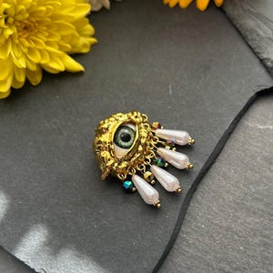 Pu&ograve; includere: Una spilla a forma di occhio dorata con un occhio verde e accenti di perle bianche. L'occhio &egrave; circondato da una cornice dorata testurizzata e le perle sono attaccate alla parte inferiore della cornice.