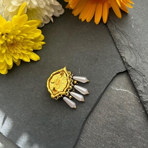 Pu&ograve; includere: Broche dorata con un designo dettagliato e perle bianche pendenti. La broche ha una chiusura a spilla sul retro.