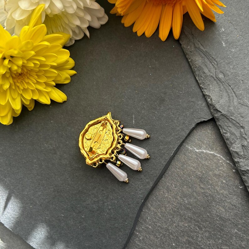 Pu&ograve; includere: Broche dorata con un designo dettagliato e perle bianche pendenti. La broche ha una chiusura a spilla sul retro.