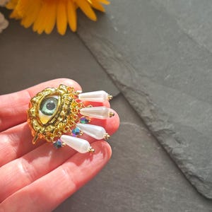 Pu&ograve; includere: Un ciondolo a forma di occhio dorato con un occhio verde e dettagli in perle bianche. Il ciondolo &egrave; appeso a una catena dorata.