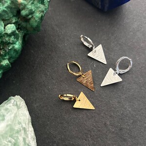 May include: Four pairs of geometric earrings with triangle pendants. Two pairs are silver, one is gold, and one is bronze. The earrings are on a dark surface with green and blue mineral accents.