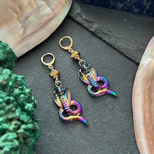 May include: A pair of iridescent cobra earrings. The earrings feature a cobra design with a rainbow metallic finish. The earrings have gold-coloured ear wires and decorative beads. The earrings are displayed on a dark grey surface.