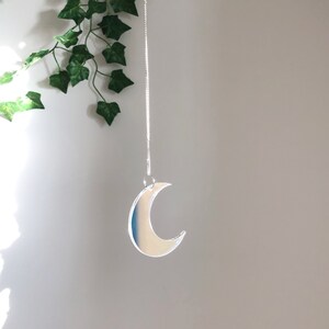 May include: A silver crescent moon shaped ornament hanging from a silver chain against a white wall.