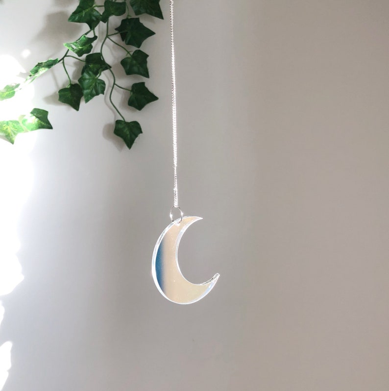 May include: A silver crescent moon shaped ornament hanging from a silver chain against a white wall.