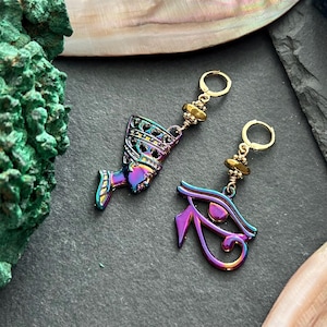 May include: A pair of iridescent earrings featuring an Egyptian theme. One earring is shaped like the bust of Nefertiti, the other is the Eye of Horus. Both have gold-colored hoops and accents.