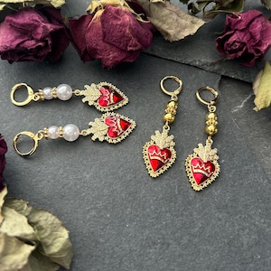 May include: Three pairs of gold earrings with red heart charms. The hearts have a gold crown and ornate detailing. One pair has white pearl accents.