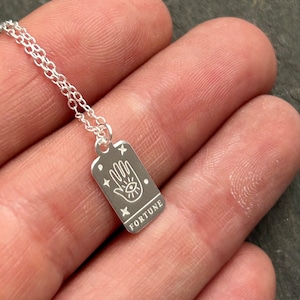 May include: Silver necklace with a rectangular pendant featuring a hand with an eye in the palm and the word "FORTUNE" inscribed below.