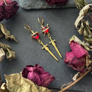 May include: A pair of gold-coloured earrings with a heart-shaped red gemstone and a sword design. The earrings have a lever back closure. The earrings are displayed on a dark surface with dried rose petals.