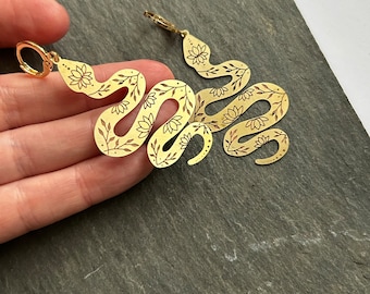 May include: A pair of gold-tone earrings shaped like snakes with floral designs. The earrings are dangling and have a hook closure.