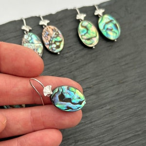 May include: A pair of silver earrings with oval-shaped abalone shell pendants. The shells have iridescent green, blue, and brown colors.
