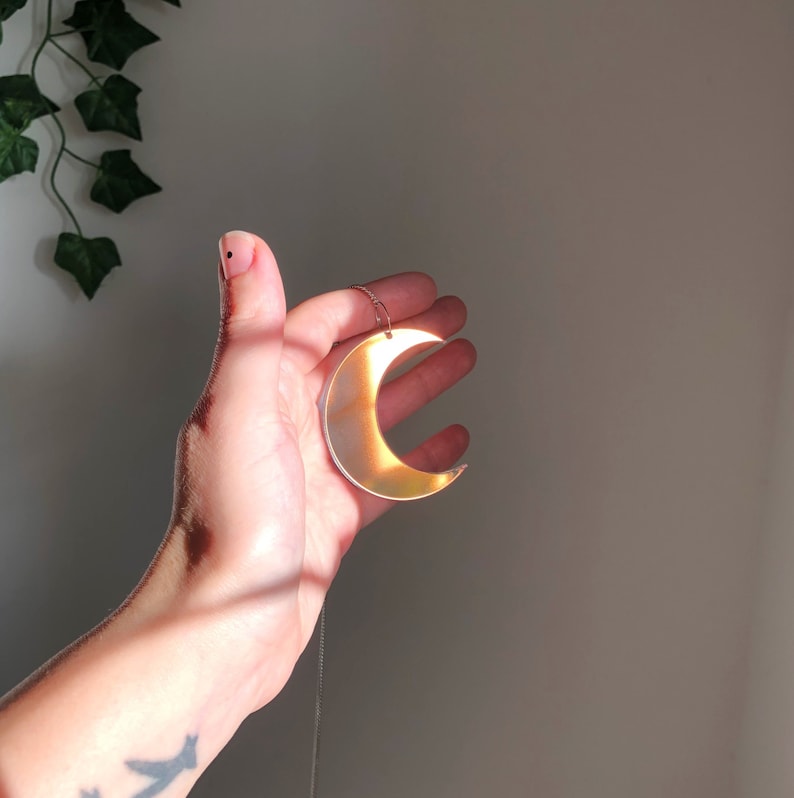 May include: A gold crescent moon pendant necklace with a iridescent finish. The necklace is hanging from a person's hand.