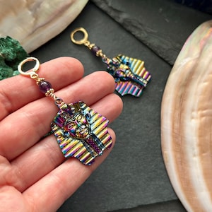 May include: A pair of iridescent earrings featuring a pharaoh design. The earrings have a gold-colored hoop and a strand of small purple beads. The pharaoh pendant is a mix of colors, including gold, purple, blue, and green.