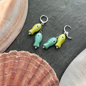 Könnte beinhalten: Ein Paar Ohrringe mit einem skurrilen Fischdesign. Jeder Ohrring hat einen silbernen Haken und eine Kette mit zwei Fischanhängern in Gelb und Türkis. Die Fischanhänger haben kleine weiße Augen und silberne Perlenakzente. Der Hintergrund ist dunkelgrau.