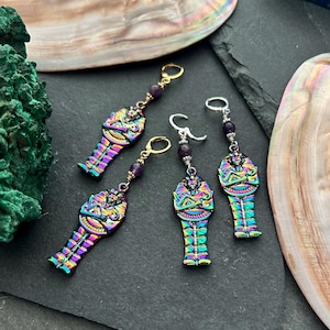 May include: Four pairs of iridescent mummy-shaped earrings. The earrings have a colourful, metallic finish with gold or silver ear wires and purple beads. The earrings are displayed on a dark surface with shell accents.