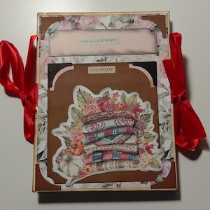 Puede incluir: Una caja decorativa con un diseño floral y temático de otoño. La caja presenta una pila de mantas, calabazas y el texto "GOOD DAY" y "Life is a JOURNEY". Cintas rojas atadas en el lateral.