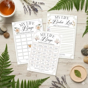 May include: Three white paper sheets with floral accents and text. The top sheet reads "MY LIFE Bucket List" and the bottom two read "MY LIFE Bingo". The bingo cards have a grid of boxes with prompts. A cup of tea and decorative elements surround the sheets.