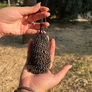 Op de afbeelding: Een kleine, donkere metalen maliënkolderzak met een leren koord, vastgehouden in de handen van een persoon. De zak is gemaakt van kleine, in elkaar grijpende metalen ringen. De achtergrond is wazig, met groene bomen en droog gras zichtbaar.