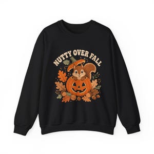 Puede incluir: Una sudadera negra de manga larga con un gráfico de una ardilla marrón sentada dentro de una calabaza naranja tallada. El diseño incluye hojas de otoño, bellotas y el texto "NUTTY OVER FALL" en una tipografía beige curvada.