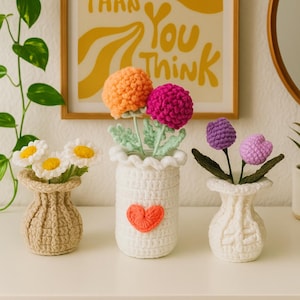 Könnte beinhalten: Drei gehäkelte Blumenarrangements in kleinen Vasen. Eine Vase enthält weiße Gänseblümchen mit gelben Zentren, eine weitere orangefarbene und fuchsienfarbene Blumen und die letzte lila Blumen. Ein gerahmter Druck mit dem Text "Maybe it is easier than you think" hängt im Hintergrund.