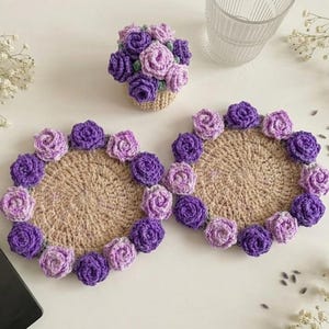 May include: Handmade crochet coasters with a floral design. The coasters are beige with a border of purple and lavender roses. A small crochet basket with purple and lavender roses is also present.