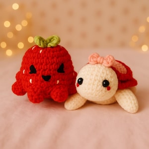 May include: Two handmade crochet toys: a red strawberry with a green stem and a white turtle with a pink bow. The strawberry has black eyes and a smiling mouth. The turtle has black eyes and a red shell.