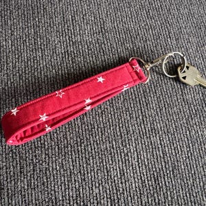 May include: A red fabric wristlet key holder with white star patterns. The key holder is attached to a silver-colored metal clasp and a key. The key holder is on a gray textured surface.