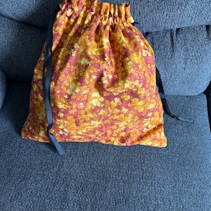 May include: A drawstring bag with a fall-themed leaf pattern in shades of orange, yellow, and red. The bag has a gathered top and is secured with black ribbon drawstrings. The bag is made of fabric and is likely used for storage or carrying items.