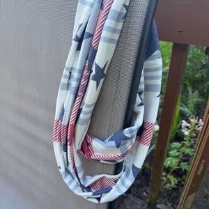 May include: An infinity scarf featuring a patriotic pattern of navy blue stars and red and white diagonal stripes on a cream-colored background. The fabric is lightweight and draped in a continuous loop, showcasing the repeating graphic design.