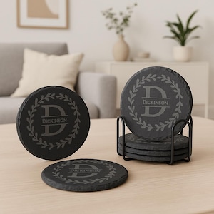 May include: A set of personalized slate coasters with a black metal holder. Each round coaster features the name "Dickinson" and the letter "D" within a laurel wreath design. The coasters are displayed on a light-colored wooden table.