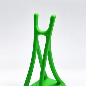 May include: A bright lime green stand featuring a square base and a vertical support structure that tapers upward into a U-shaped cradle at the top. The object has a smooth, matte finish and a symmetrical design with open spaces between the supports.