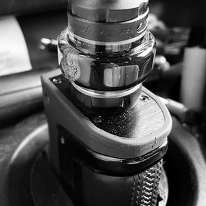 May include: Black and silver vaping device with a cylindrical top and a rectangular base. The device has a clear glass component and a textured grip. The brand name "Geek Vape" is visible on the device.