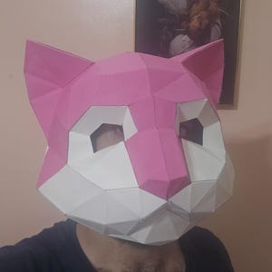 Op de afbeelding: Een roze en wit geometrisch kattenmasker. Het masker is gemaakt van gevouwen papier of karton, met een polygonale vormgeving. De oren, neus en voorhoofd zijn roze, terwijl het gezicht wit is. Het masker bedekt het hele gezicht.