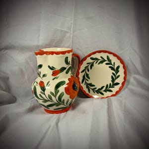 May include: A cream-colored ceramic pitcher and plate set. The pitcher features orange poppy flower designs with green leaves and a red scalloped rim. The plate has a green leaf design and a red scalloped edge. The set is likely for serving.
