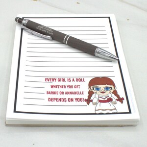 May include: A notepad with lined paper and a dark brown pen. The notepad has the text "EVERY GIRL IS A DOLL WHETHER YOU GET BARBIE OR ANNABELLE DEPENDS ON YOU!" and a cartoon doll illustration.